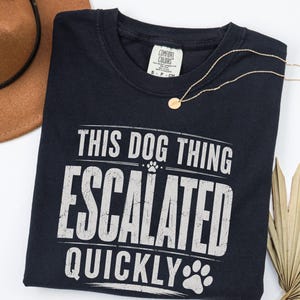 Puede incluir: Una camiseta de manga corta azul marino oscuro con el texto blanco desgastado "THIS DOG THING ESCALATED QUICKLY" y pequeños gráficos de huellas. La camiseta tiene un cuello redondo y una etiqueta de la marca Comfort Colors en el interior.