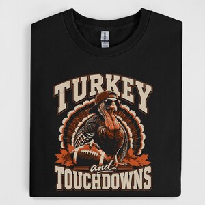 Könnte beinhalten: Ein schwarzes T-Shirt mit einer Grafik eines Truthahns, der eine umgedrehte Baseballkappe und eine Sonnenbrille trägt und einen Football hält. Der Text "TURKEY and TOUCHDOWNS" ist in einer fetten, abgenutzten Schriftart aufgedruckt.