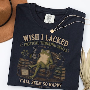 May include: A dark navy blue t-shirt featuring a graphic of a green frog sitting in an armchair, holding a mug labelled "Over Thinking Club." The text reads "Wish I Lacked Critical Thinking Skills" and "Y'all Seem So Happy" with stacks of books.
