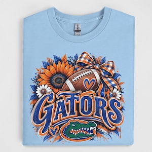 May include: A light blue t-shirt featuring a graphic print of a football, a large sunflower, white daisies, and a plaid bow. The word GATORS is printed in large blue and orange letters above a green alligator head icon with an orange outline.
