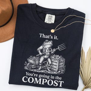 May include: A black t-shirt featuring a graphic of a frog holding a pitchfork next to a wheelbarrow and a crate of vegetables. The text reads "That's it. You're going in the COMPOST". The shirt has a Comfort Colors brand label at the neck.