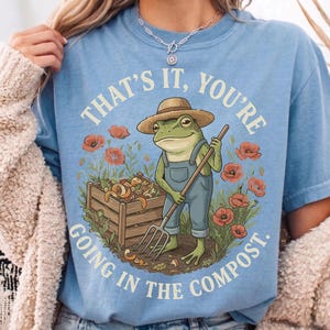 Op de afbeelding: Een lichtblauw t-shirt met een afbeelding van een groene kikker met een strooien hoed en een blauwe tuinbroek, die een riek vasthoudt naast een houten compostbak. Rode klaprozen omringen de scène met de tekst THAT'S IT, YOU'RE GOING IN THE COMPOST.
