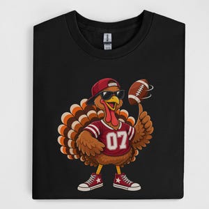 May include: A black t-shirt featuring a graphic of a brown turkey wearing a red baseball cap, black sunglasses, and a red jersey with the number 07. The turkey is spinning a brown football on its finger and wearing red and white trainers.
