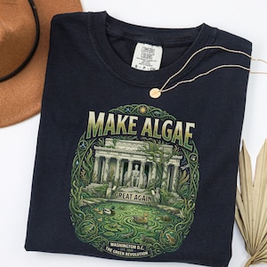 May include: A dark navy blue t-shirt featuring a graphic print of the Lincoln Memorial overgrown with green algae and vines. The text reads "MAKE ALGAE GREAT AGAIN" above the monument and "WASHINGTON D.C. EST. 2026 THE GREEN REVOLUTION" below.