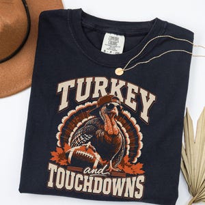 Könnte beinhalten: Ein dunkelblaues T-Shirt mit einer Grafik eines Truthahns, der eine Baseballkappe und eine Sonnenbrille trägt und einen Football hält. Der Text TURKEY und TOUCHDOWNS ist in einer hellen, fetten Schriftart aufgedruckt.