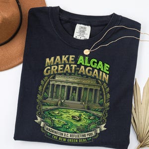 May include: A dark navy blue t-shirt featuring a graphic of the Lincoln Memorial with a green algae-filled reflecting pool. The text reads "MAKE ALGAE GREAT AGAIN" and "WASHINGTON D.C. REFLECTING POOL THE NEW GREEN DEAL" in green and gold lettering.