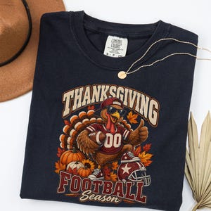 May include: A dark navy blue t-shirt featuring a graphic of a turkey wearing a baseball cap, sunglasses, and a football jersey. The turkey holds a football, surrounded by pumpkins and autumn leaves. Text reads "THANKSGIVING FOOTBALL SEASON".