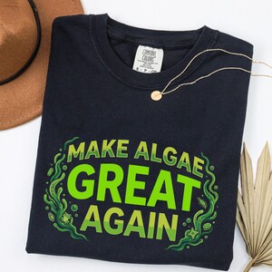 May include: A dark navy blue t-shirt featuring the text "MAKE ALGAE GREAT AGAIN" in bright green letters. The design is surrounded by green swirling algae patterns and a small frog illustration. The shirt has a Comfort Colors brand neck label.