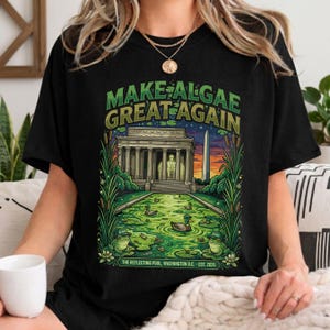 May include: A black t-shirt featuring a graphic print of the Lincoln Memorial and Washington Monument with a green algae-filled reflecting pool, ducks, and frogs. The text reads "MAKE ALGAE GREAT AGAIN" and "THE REFLECTING POOL, WASHINGTON D.C. - EST. 2026".
