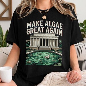 Könnte beinhalten: Ein schwarzes T-Shirt mit einem Grafikdruck des Lincoln Memorial, umgeben von einem grünen, algenbedeckten Teich mit Fröschen und einer Ente. Der Text lautet MAKE ALGAE GREAT AGAIN, ergänzt durch ALGAE NATION und EST 2026.