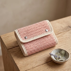 May include: A rectangular crochet wallet featuring a dusty pink textured body with a cream-colored border. The flap is secured by two silver-toned metal snap buttons. The wallet has a structured, folded design with visible yarn stitches throughout.