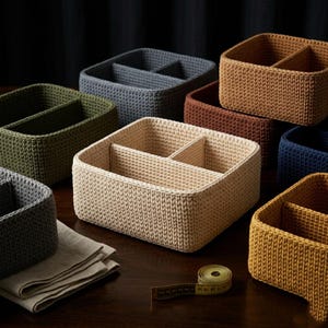 K&ouml;nnte beinhalten: Eine Sammlung gewebter Aufbewahrungsk&ouml;rbe in verschiedenen Farben, darunter Beige, Gr&uuml;n, Blau und Braun. Jeder Korb ist in zwei F&auml;cher unterteilt. Ein Ma&szlig;band und ein gefaltetes Leinentuch sind ebenfalls zu sehen.