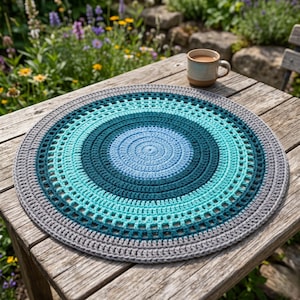 Puede incluir: Un salvamanteles redondo de ganchillo en tonos azules, turquesas y grises. El diseño circular presenta anillos concéntricos de color, con un patrón texturizado. Una taza de café está sobre una mesa de madera.