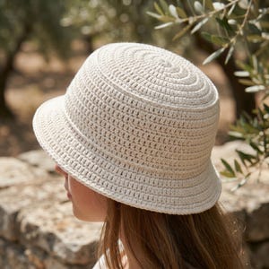 Può includere: Un cappello a secchiello all'uncinetto color crema. Il cappello presenta una corona arrotondata con cerchi concentrici e una tesa leggermente inclinata verso il basso. Il cappello è realizzato in un materiale tessuto strutturato.