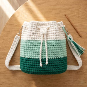 Könnte beinhalten: Eine gehäkelte Beuteltasche mit einem dreifarbigen Ombré-Design in Weiß, hellem Minzgrün und dunklem Waldgrün. Die Tasche hat einen weißen Kordelzug, einen passenden weißen Häkel-Schulterriemen und eine dekorative Quaste in Grün und Weiß.