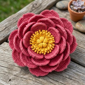 May include: A handcrafted crochet flower featuring multiple layers of deep pink, rounded petals arranged in a circular pattern. The center of the flower is composed of numerous small, bright yellow, textured yarn loops that create a dense, raised core.