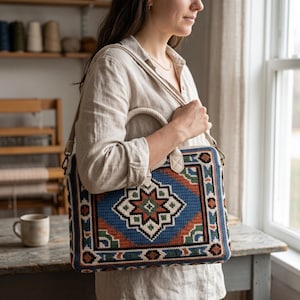 May include: A rectangular crochet bag featuring a geometric star pattern in blue, orange, green, and cream. The bag has a thick, braided cream-colored handle and a matching detachable shoulder strap with metal hardware clips on each side.