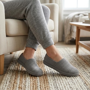 May include: A pair of grey slip-on slippers featuring a textured, crocheted knit construction with a rounded toe and a low-profile design. The slippers are worn with matching grey knit joggers, showcasing a consistent color and fabric aesthetic.