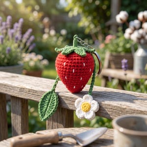 May include: A red crocheted strawberry keychain with white seed details, green leaves, and a white and yellow flower charm. The keychain is attached to a silver metal ring. The strawberry is sitting on a wooden surface.