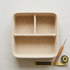 K&ouml;nnte beinhalten: Ein quadratischer, cremefarbener H&auml;kelkorb mit zwei Innenteilern. Ein H&auml;kelhaken und ein gelbes Ma&szlig;band liegen neben dem Korb. Der Korb ist handgefertigt und eignet sich zum Ordnen kleiner Gegenst&auml;nde.