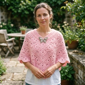 May include: A light pink, crocheted poncho with a V-neck and scalloped edges. A silver butterfly brooch adorns the neckline. The poncho is worn over a white shirt, with a garden setting in the background.