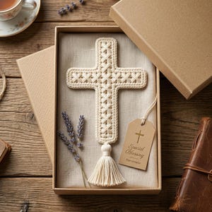 Op de afbeelding: Een crèmekleurig gehaakt kruis met een decoratieve kwast aan de onderkant, gepresenteerd in een kartonnen geschenkdoos. Een klein rechthoekig label bevestigd met een touwtje toont een gouden kruisicoon en de tekst Special Blessing Handmade.