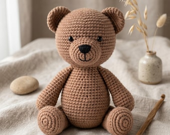 May include: A handmade brown crochet teddy bear with a black nose and eyes. The bear is sitting on a neutral-colored surface. A small vase with dried flowers and a crochet hook are in the background.