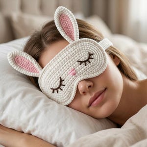 Op de afbeelding: Een wit gehaakt slaapmasker met twee lange konijnenoren met roze centra. Het masker heeft geborduurde gesloten ogen met donkerbruine wimpers en een kleine roze gestikte neus. Het ontwerp bevat een bijpassende witte gehaakte band voor bevestiging.