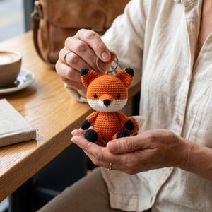 Op de afbeelding: Een gehaakte vossen sleutelhanger in oranje, wit en zwart. De vos heeft een rond lichaam, kleine oren en een witte snuit. De sleutelhanger wordt door een persoon vastgehouden. De achtergrond omvat een houten tafel, een koffiekopje en een leren tas.