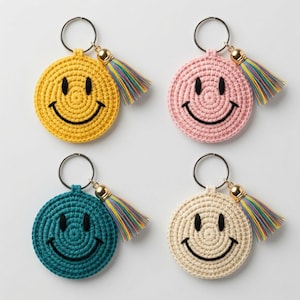 May include: Four circular crochet keychains featuring a smiley face design with black stitched eyes and a curved mouth. Each keychain includes a silver-toned metal ring and a multi-coloured tassel in yellow, pink, teal, and cream yarn colours.