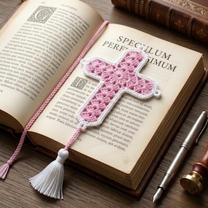 Pode incluir: Um marcador de página de crochê em formato de cruz, apresentando um padrão floral rosa claro com uma borda branca. O marcador inclui um longo cordão trançado rosa claro que se estende do topo à base, terminando em uma borla branca.