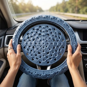 May include: A dusty blue crochet steering wheel cover featuring a circular mandala-style pattern in the center and a textured shell stitch design around the outer rim, fitted securely onto a car steering wheel with a dark interior dashboard.