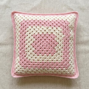 May include: Square crochet pillow in a granny square pattern. The pillow features alternating bands of light pink and cream-colored yarn, with a solid pink border. The handmade pillow is a decorative item for a bed or sofa.