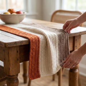 May include: A handmade, striped table runner in shades of orange, cream, and brown. The runner is made of a textured, crocheted material and is draped over a wooden table. The runner is approximately 60 inches long.