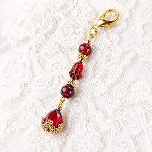 May include: A gold-colored charm with a lobster clasp, featuring red glass beads and a teardrop-shaped pendant with a gold filigree base. The charm has a floral design and is likely used for jewelry or accessories.
