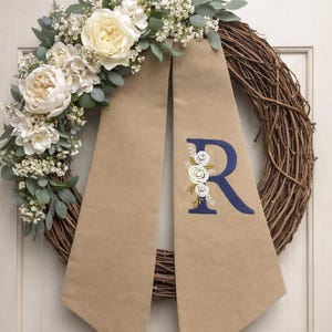 May include: A grapevine wreath adorned with cream-colored flowers, greenery, and a tan fabric sash. The sash features a navy blue embroidered letter "R" with floral accents. This decorative wreath is perfect for adding a touch of elegance to any home.