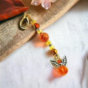 May include: A gold charm with an angel wing design and orange and yellow glass beads. The charm is attached to a gold clasp.