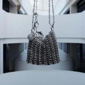 Può includere: Una borsa a mano color argento realizzata con linguette di lattine metalliche intrecciate, dotata di una lunga tracolla a catena e di un ciondolo decorativo composto da due sfere glitterate e una foglia metallica sul lato.