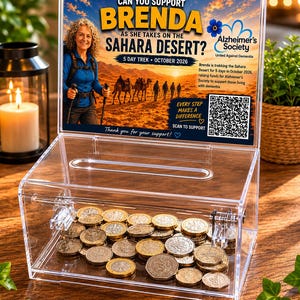 May include: A clear acrylic donation box containing coins. The sign reads "Can You Support Brenda as She Takes on the Sahara Desert?" with a photo of a woman hiking. The sign also includes information about the Alzheimer's Society and a QR code.