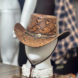 Könnte beinhalten: Ein Cowboyhut mit braun-schwarzem Schlangenledermuster, ausgestattet mit einem schwarzen Lederband und einem mittigen, goldfarbenen runden Emblem. Der Hut verfügt über ein braunes Kinnband mit schwarzem Verstellclip sowie eine geschwungene Krempe.
