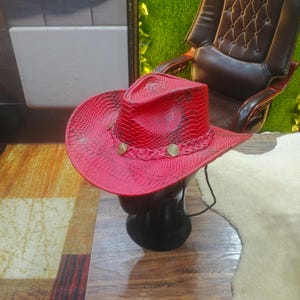Può includere: Un cappello da cowboy rosso brillante caratterizzato da un motivo a pelle di serpente testurizzato sulla corona e sulla tesa. Il cappello è decorato con una fascia intrecciata rossa e tre borchie metalliche circolari color oro. Un sottogola nero pende.