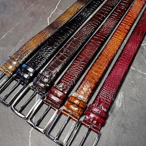 Pode incluir: Uma coleção de seis cintos com um padrão texturizado de imitação de crocodilo. Os cintos estão disponíveis em várias cores, incluindo castanho claro, preto, castanho escuro, castanho avermelhado, castanho médio e vermelho escuro, cada um com uma fivela de metal prateado.