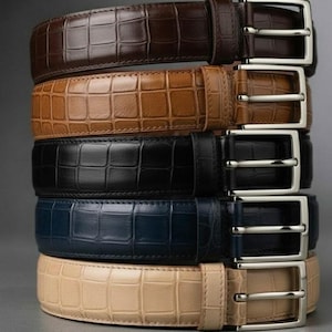 Könnte beinhalten: Ein Stapel aus fünf Gürteln mit Krokodilprägung. Die Gürtel sind in den Farben Dunkelbraun, Hellbraun, Schwarz, Marineblau und Beige gehalten. Jeder Gürtel verfügt über eine silberfarbene Metallschnalle mit Dorn und eine passende Schlaufe.