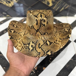 Pode incluir: Um chapéu de cowboy com um padrão de pele de cobra em tons de bege e preto em toda a copa e na aba larga e curva. O chapéu tem uma forma estruturada com o topo vincado e um vinco definido ao longo do centro da copa.