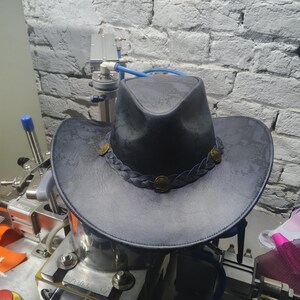 May include: A dark gray cowboy hat with a braided band and decorative gold-colored buttons. The hat has a wide brim and a textured appearance, suggesting a leather or similar material. The hat is a classic Western style.