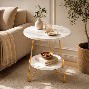 Peut inclure: Une table d'appoint ronde avec deux plateaux en faux marbre blanc et trois pieds fins en métal doré. Le plateau supérieur supporte un vase en céramique blanche avec des feuilles vertes, un bougeoir en verre et un petit plat décoratif.