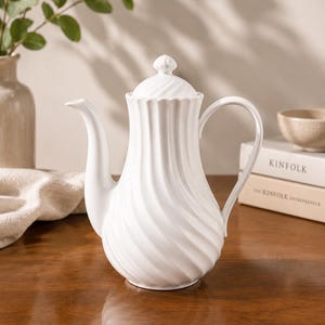 Könnte beinhalten: Eine weiße Keramik-Teekanne mit einem Spiraldesign und einem passenden Deckel. Die Teekanne hat einen gebogenen Ausguss und Griff. Der Hintergrund umfasst eine Vase mit Grün, Bücher und einen Holztisch.
