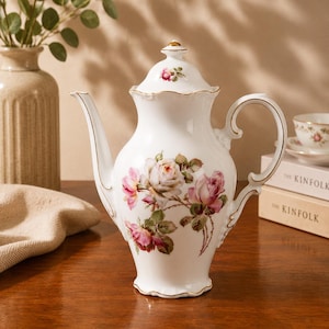 Könnte beinhalten: Weiße Porzellan-Teekanne mit einem floralen Rosenmuster in Rosa und Creme, akzentuiert mit goldenen Verzierungen. Die Teekanne hat einen gebogenen Ausguss und Henkel sowie einen passenden Deckel. Eine Tasse und Untertasse sind im Hintergrund zu sehen.