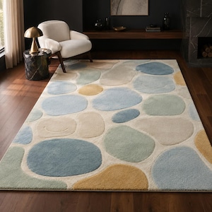 May include: A rectangular area rug featuring an abstract pattern of rounded, pebble-like shapes in shades of blue, sage green, mustard yellow, and cream. The rug has a textured, tufted surface and covers a dark wooden floor in a room.