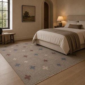 Puede incluir: Una alfombra rectangular con una base de color beige neutro y un patrón repetitivo de pequeñas formas de cruz espaciadas uniformemente en tonos apagados de rojo, azul y blanco roto. La alfombra tiene flecos cortos en los bordes.